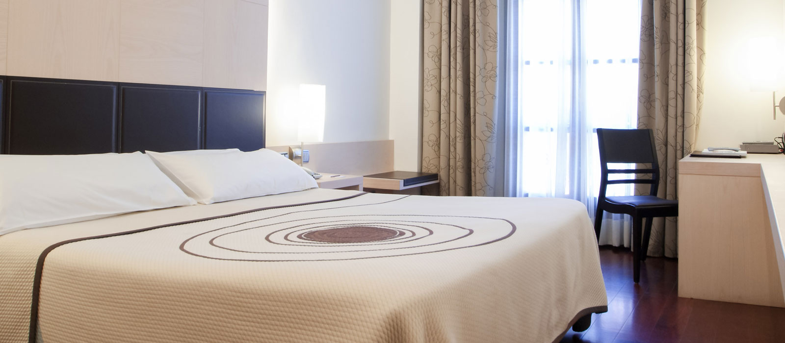 Habitación doble del Hotel Mozart en Valladolid con una gran cama, escritorio de madera clara, televisión y grandes ventanales con cortinas.