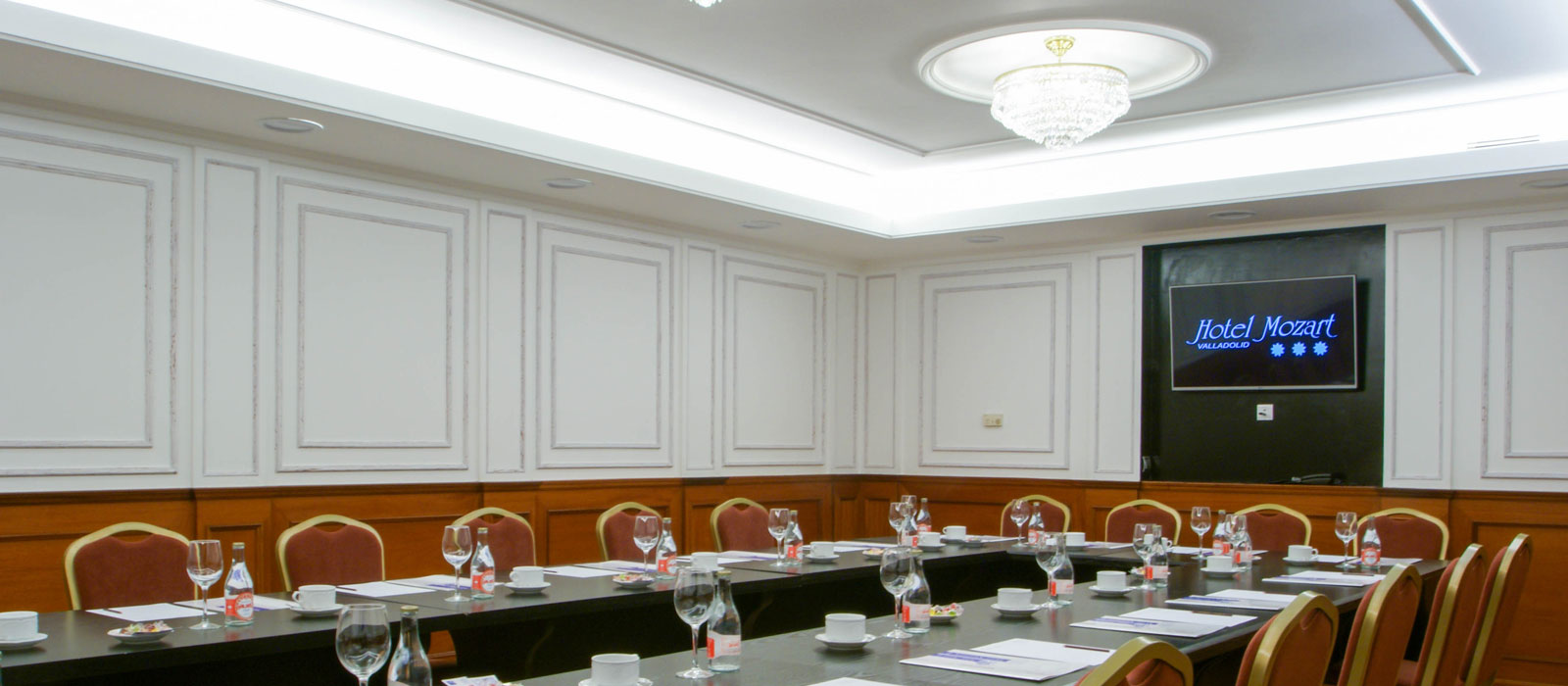 Sala de reuniones del Hotel Mozart configurada en forma de U, con mesas preparadas con agua y copas, y una pantalla mostrando el logo del hotel.